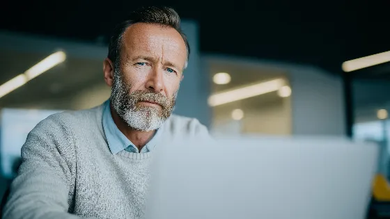 a middle-aged man with a beard in business casual clothing sits in front of a computer. His expression is thoughtful and contemplative.